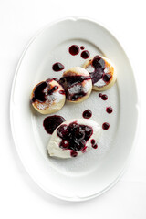 A beautifully presented dessert featuring three fluffy pancakes drizzled with dark berry sauce, accompanied by a dollop of whipped cream on a pristine white plate. Perfect for culinary showcases.