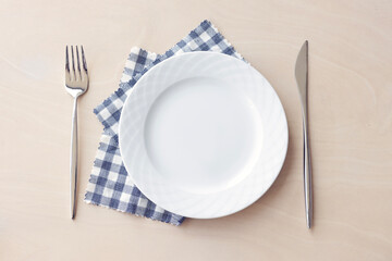 Empty white plate with blue fabric napkin, a fork and a knife on wooden table. Top view.	