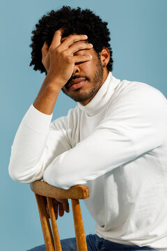 A man sitting on a wooden stool in a white turtleneck covering his fac
