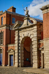 architectural shot of the Bedford Tower Gate at Dublin Castle, Ireland. The image showcases the intricate stonework of the archway, flanked by historic red brick buildings, under a clear blue sky. 