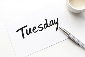The text “Tuesday” is written in black letters on a piece of paper on the table. Signboard, calendar. Advertising banner