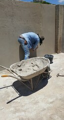 Pedreiro no trabalho de construção do muro de cimento, com carrinho de massa de cimento ao lado.