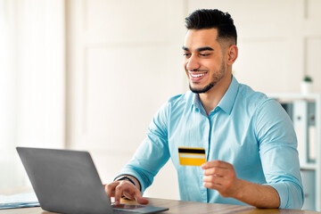 Cheerful Arab guy with laptop computer and credit card purchasing via internet at office. Smiling Eastern man buying goods online in web store while sitting at workplace