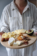 A woman holding a wooden platter with a variety of cheeses, nuts, dried fruits, and half an avocado