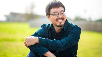 Outdoor portrait of a smiling Asian man wearing glasses and dark teal shirt sitting in a park