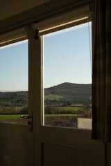 A peaceful countryside view seen through a window, featuring rolling green hills, scattered trees, a winding road, and a hill in the background, framed by the window and a partially drawn curtain.