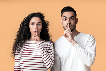 Young arab man and woman showing shh gesture at camera, mysterious middle eastern couple keeping forefingers on lips, making silence sign while standing together over yellow background, copy space