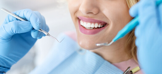 Closeup of female patient showing her beautiful white teeth while having treatment at dental clinic, dentist hands in rubber gloves holding dental tools, panorama. Woman having regular checkup