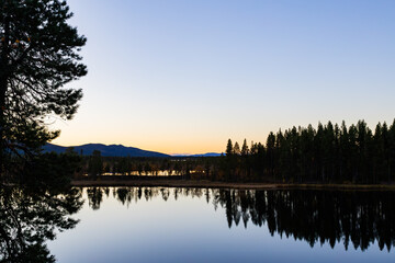 A peaceful sunset over a lake in Lapland, Sweden, with the reflection of trees in the still water.