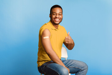 Vaccination Against Coronavirus. Happy Vaccinated African American Guy Gesturing Thumbs Up Showing Arm With Plaster After Covid-19 Vaccine Injection Over Blue Background. Corona Virus Immunization © Prostock-studio