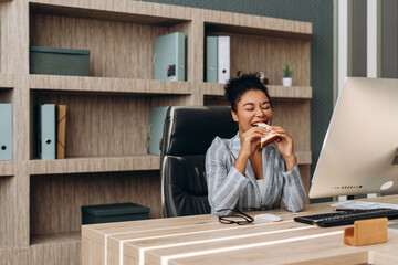 Happy businesswoman eating sandwich at office desk during lunch break