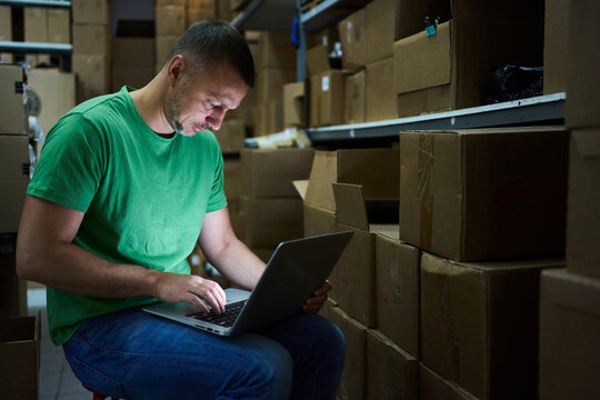 A man working on a laptop amidst cardboard boxes in a storage area