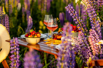 Simple countryside beautiful wedding decor, surprise romantic date outdoors, garden arrangement with lupins flowers on a meadow. Fruits, rose wine, burning candles. Sunset, candlelight, golden hour.