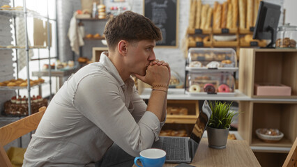 Young man sitting thoughtfully in bakery with laptop and coffee, surrounded by pastries and bread, indoors, handsome caucasian adult pondering work