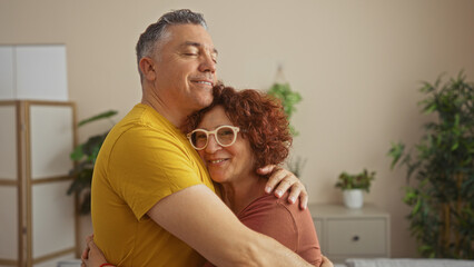 Fototapeta premium Happy middle-aged couple hugging at home in a cozy living room, both smiling and wearing casual clothes, with plants and modern decor in the background