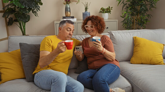 Middle-aged couple enjoying coffee and looking at a smartphone together in their cozy living room with plants, yellow pillows, and modern decor, conveying a warm, homey atmosphere - Powered by Adobe