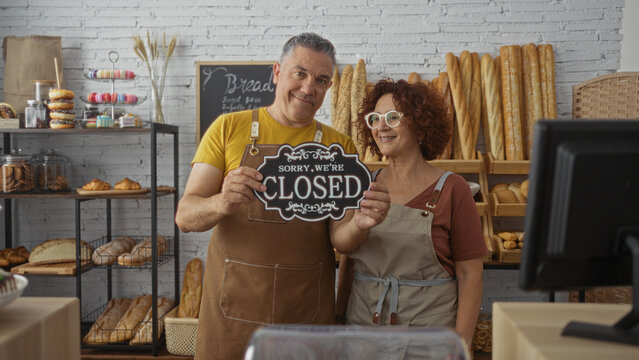 Man and woman bakers holding a closed sign in a bakery shop with various breads and pastries behind them