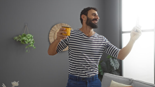 Young man enjoying coffee indoors at home while taking a selfie in the living room with a smile