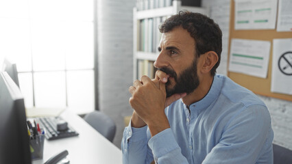 Fototapeta premium Hispanic man with a beard thinking deeply in an office room setting, characterized by a modern interior and large windows.