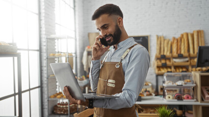 Handsome young hispanic man with a beard using a laptop and phone while working in a bakery shop...