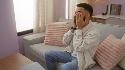 Young hispanic man with beard sitting on a couch in a cozy living room at home, looking stressed with hands covering his face