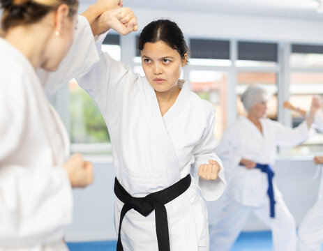 Experienced young Asian female karateka in kimono with black belt engaging kumite with rival, demonstrating martial art techniques in training room..