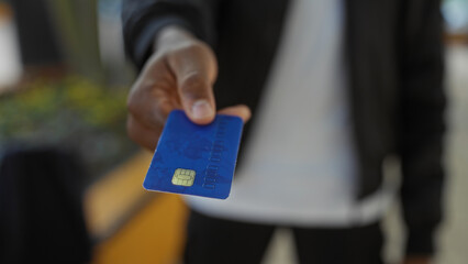 Young man in city park holds blue credit card forward against urban backdrop, highlighting modern financial transaction in outdoor setting.