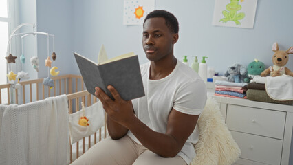 Young man reading book in bright bedroom beside cradle surrounded by stuffed animals and baby supplies depicting serene indoor home atmosphere