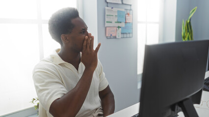 Young man in an office whispers a secret while looking at a computer screen, surrounded by a well-lit interior with documents on a board behind him