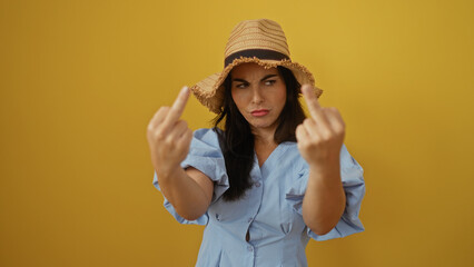 Young woman in a blue dress and straw hat making offensive gesture isolated over yellow background...