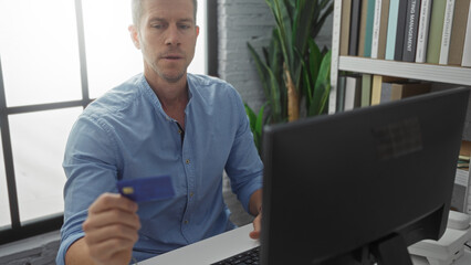 Man holding credit card in modern office setting at computer with bookshelf and plants, displaying...