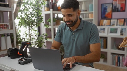 Fototapeta premium Young man working on laptop in a home decor store surrounded by shelves and plants, focused on his task in a bright, modern setting