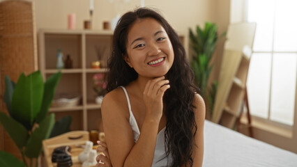 Beautiful young chinese woman smiling at spa wellness center, surrounded by indoor plants and spa essentials, creating a serene and relaxing atmosphere.