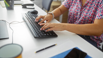 Woman typing on a keyboard at an office desk, wearing a colorful blouse, indicating a work...