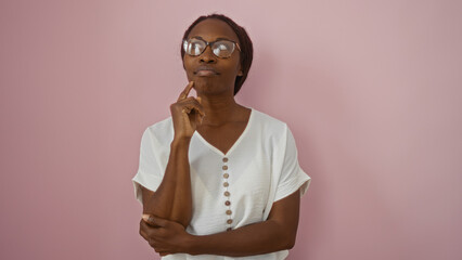 Young african american woman in white shirt standing over isolated pink background reflecting thoughtfully with a hand on her chin and the other arm crossed, wearing glasses.