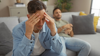 Man stressed at home with his partner relaxing on the sofa in the background, illustrating family...