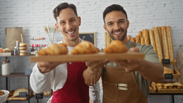 Two men wearing aprons smiling while presenting croissants in a bakery shop with various baked goods in the background