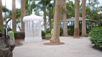 Tropical resort scene with blurred background featuring palm trees, defocused white gazebo surrounded by lush greenery and a path lined with natural stones.