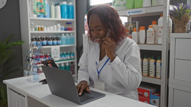 Woman pharmacist talking on phone and using laptop in modern pharmacy store with shelves full of medical products and eyeglasses display