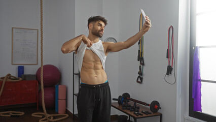 Hispanic man with a beard taking a selfie while showing abs in a gymnasium with various fitness...