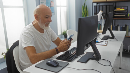 Man with grey hair and beard in a workplace, using a smartphone at his desk, surrounded by office equipment and files.