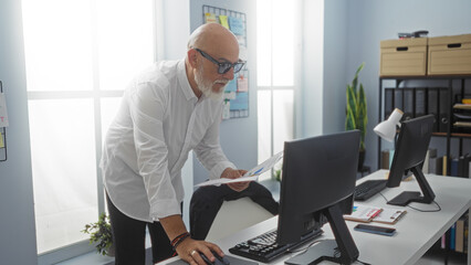 Handsome senior man with a beard analyzing documents in an office environment, standing near a desk with a computer and bright indoor lighting.