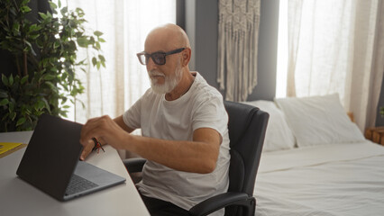 Mature man with grey hair and beard working on a laptop in a modern bedroom with a bed and indoor plant.
