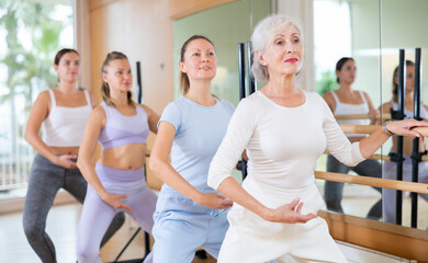 Slim aged woman and other attendees practicing ballet at ballet barre in dance studio during...