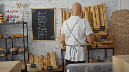 Mature man with bald head in bakery facing bread display, wearing apron, inside well-lit shop with...