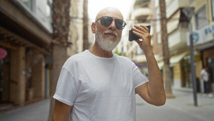 Mature man with grey hair and beard listening to a voice message on his phone while standing...
