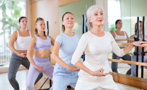 Slim aged woman and other attendees practicing ballet at ballet barre in dance studio during training session