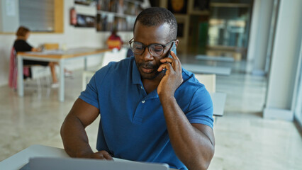 Man in office talks on phone, focused on laptop, in an open, modern workspace filled with natural...