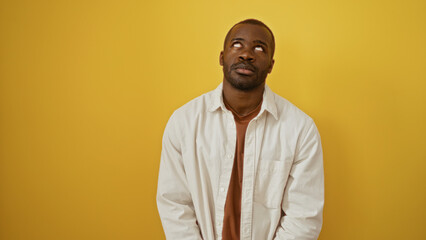 Handsome young african american man standing over isolated yellow background, looking up with a contemplative expression