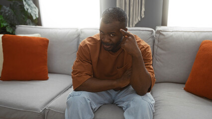 Obraz premium Young, attractive african american man sitting thoughtfully on a gray couch in a cozy living room with orange pillows, implying a relaxed indoor setting at home.